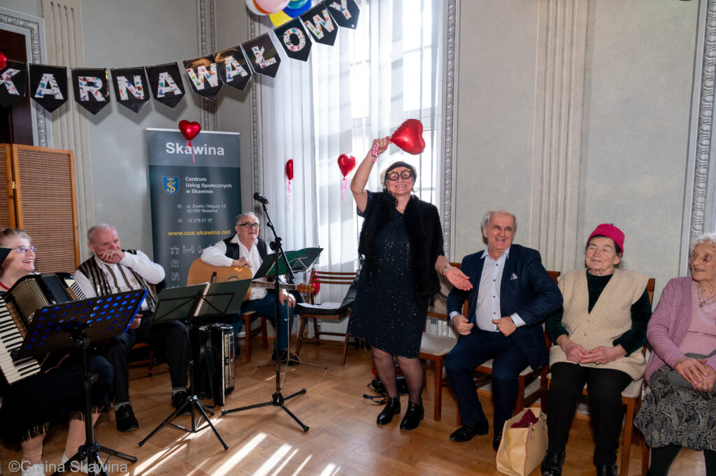 Po lewej stronie jest kapela w środkowej częśći stoi seniorka która w ręce trzyma balon w kształcie czerwonego serca, po prawej stronie siedzą seniorzy 