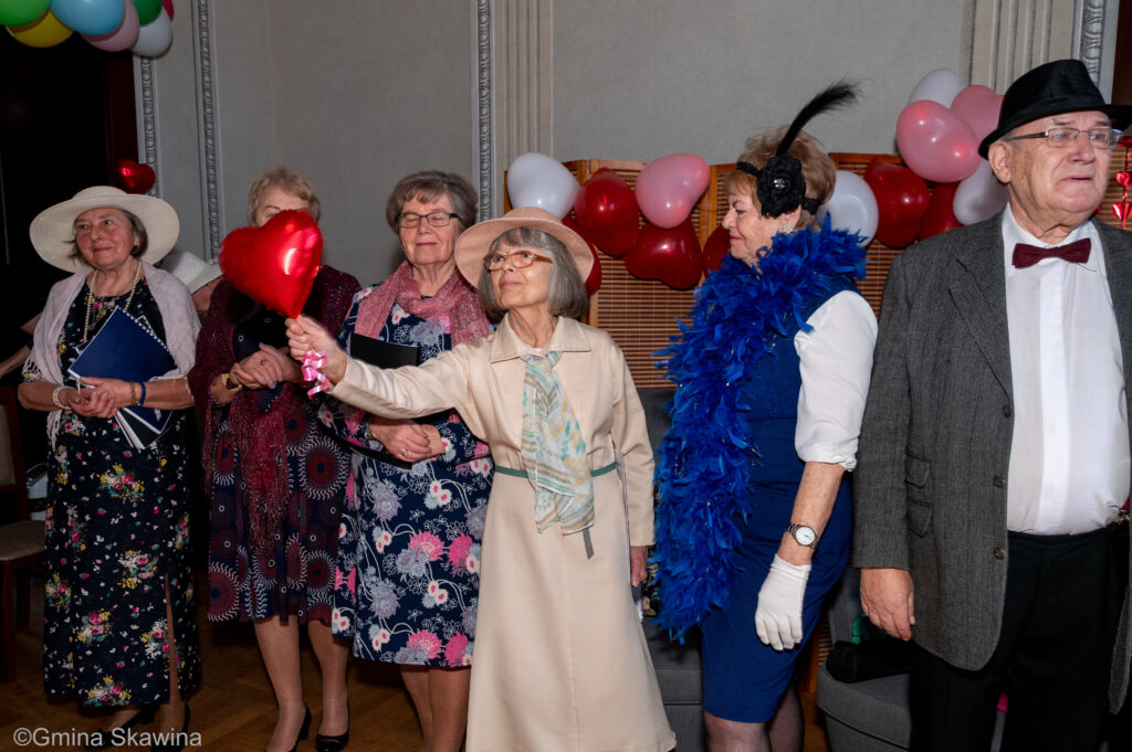 Grupa seniorów, seniorka w samym środku grupy trzyma balon w kształcie czerwonego serca 
