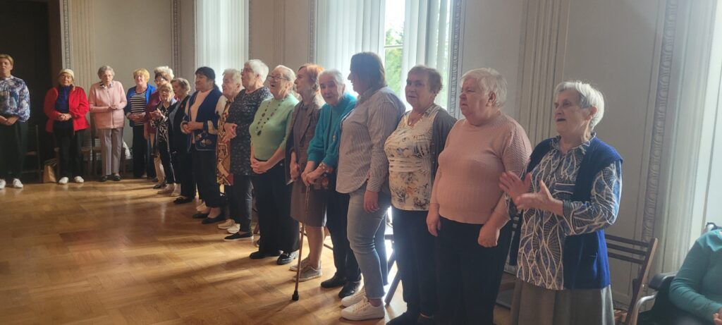 Seniorzy stoją w półkole i śpiewają