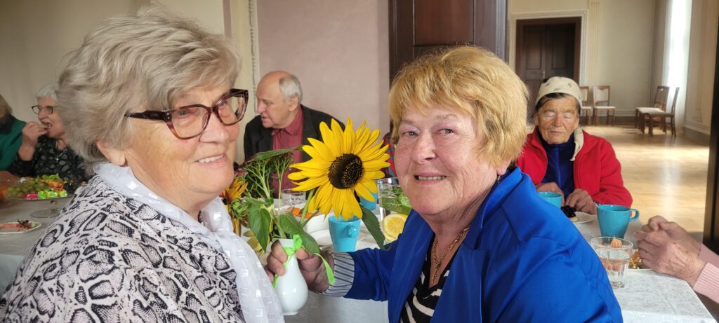 Na pierwszym planie są uśmiechnięte seniorki , jedna z nich trzyma w ręku wazon z kwiatami. Za nimi siedzą seniorzy