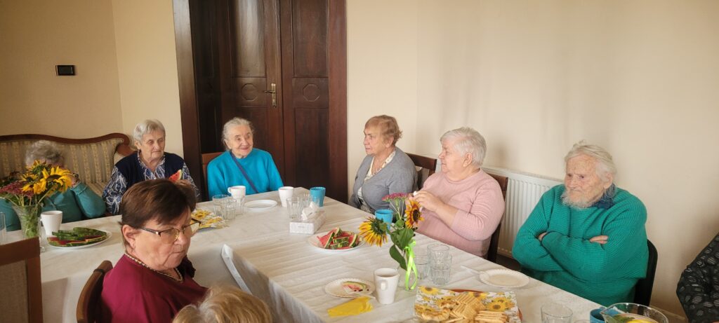 Seniorzy siedzą i rozmawiają wokół stolika na którym są przekąski