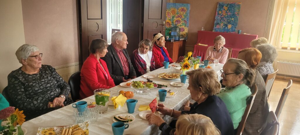 Seniorzy siedzą i rozmawiają wokół stolika na którym są przekąski