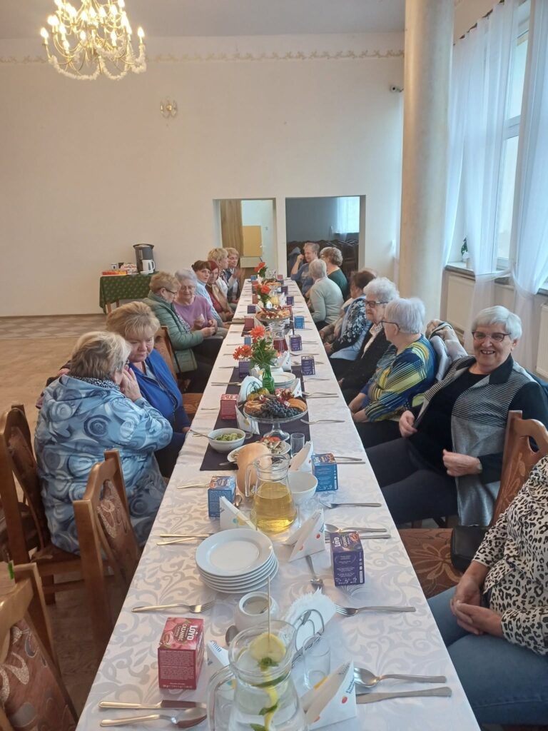 Uśmiechnięci Seniorzy siedzą przy wspólnym stole zastawionym jedzeniem i rozmawiają ze sobą