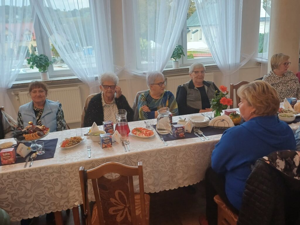 Uśmiechnięci Seniorzy siedzą pzey wspólnym stole zastawionym jedzeniem