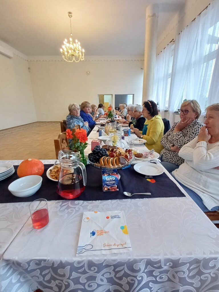 Seniorzy siedzą przy wspólnym stole zastawionym jedzeniem