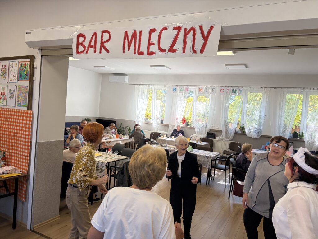 Grupa seniorek tańczy pod banerem z napisem Bar mleczny