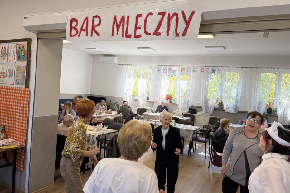 Grupa seniorek tańczy pod banerem z napisem Bar mleczny