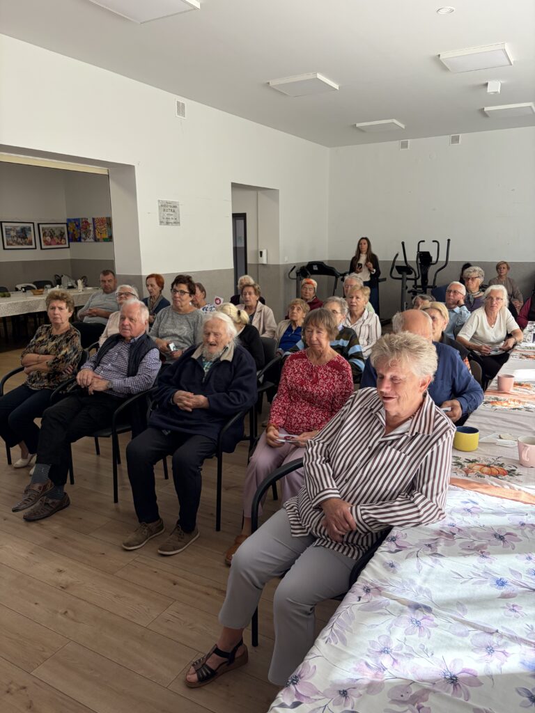Seniorzy siedzą w równych rzędach krzesełek na sali
