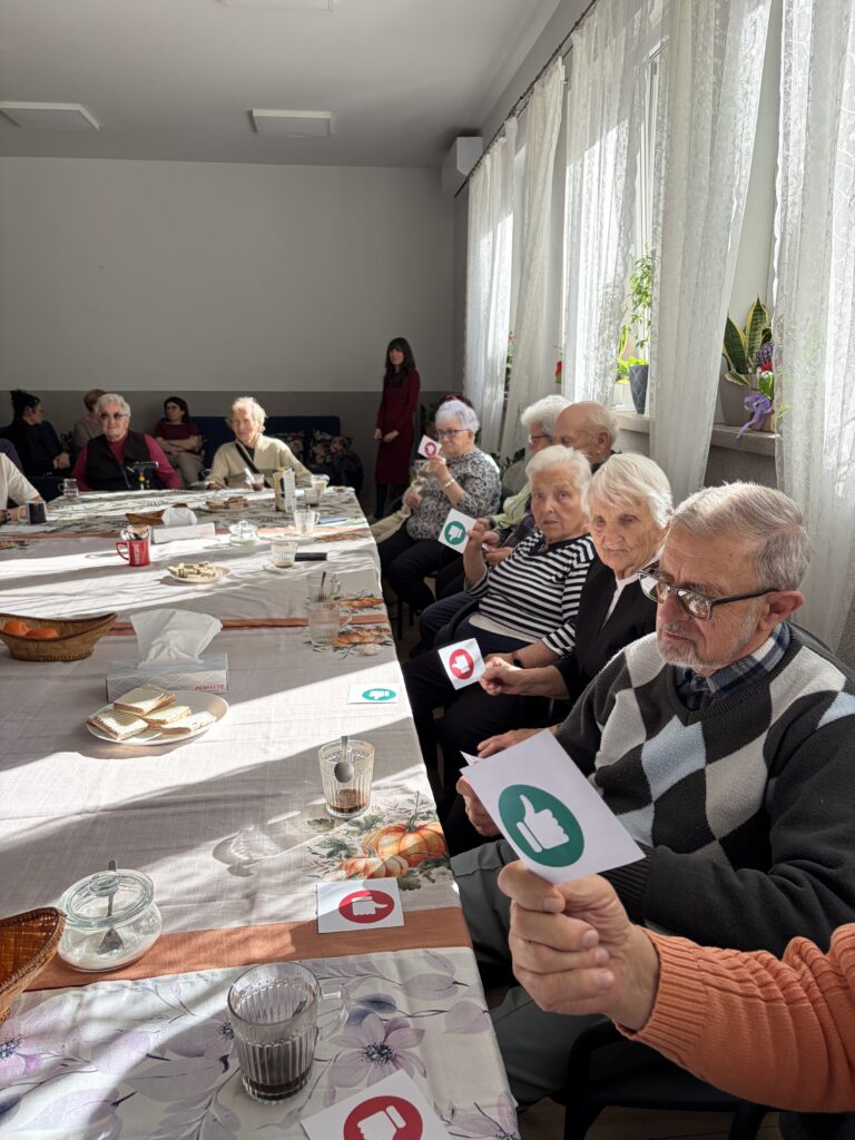 Seniorzy siedzą przy wspólnym stole i w rękach trzymają karteczki z kciukiem do góry lub na dół