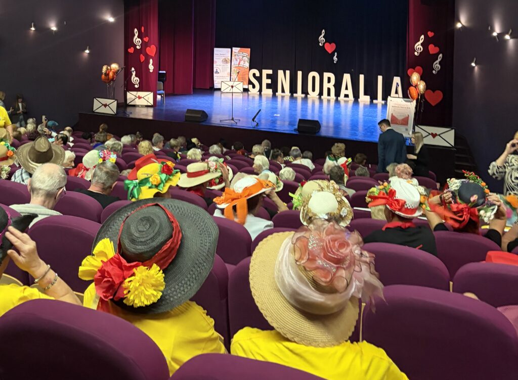 Aula, osoby siedzące na widowni mają kolorowe, ozdobione kwiatami kapelusze na głowach. Scena pięknie oświetlona na której jest duży napis Senioralia