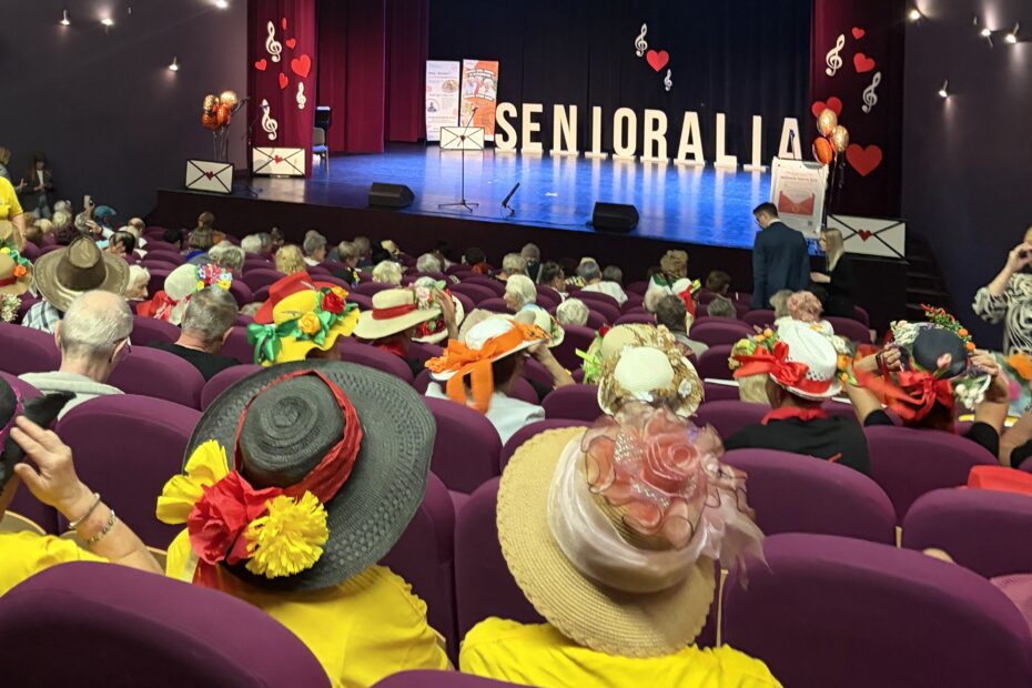 Aula, osoby siedzące na widowni mają kolorowe, ozdobione kwiatami kapelusze na głowach. Scena pięknie oświetlona na której jest duży napis Senioralia