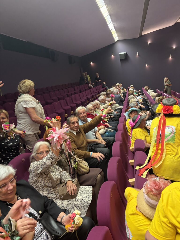 Grupa seniorów siedzi w auli i pokazuje ręcznie robione kwiaty, które trzymają w dłoniach