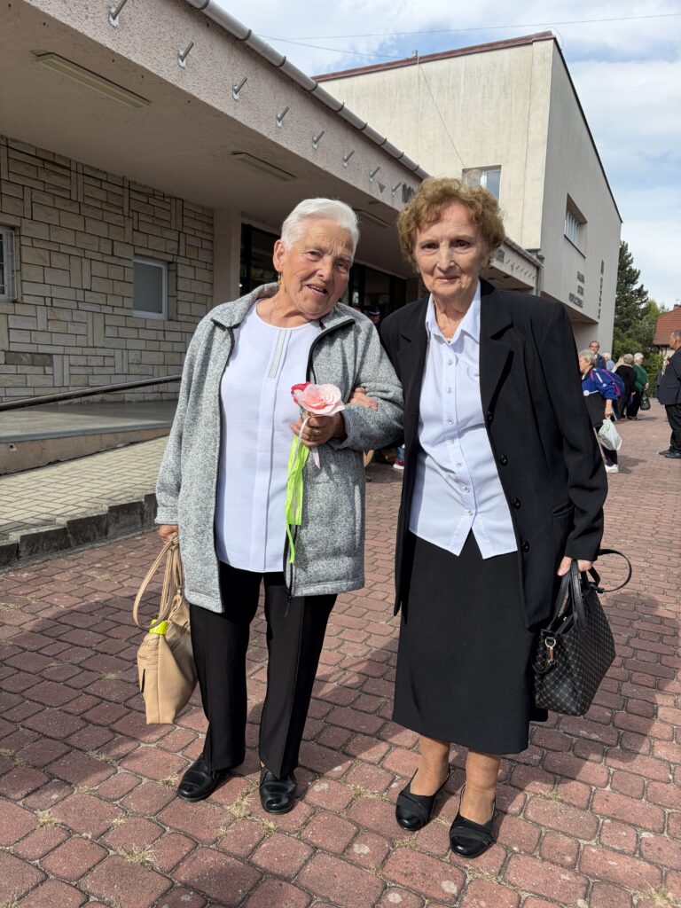 Dwie uśmiechnięte seniorki stoją przed budynkiem. Pani po prawej tronie trzyma w dłoni kwiat zrobiony z bibuły