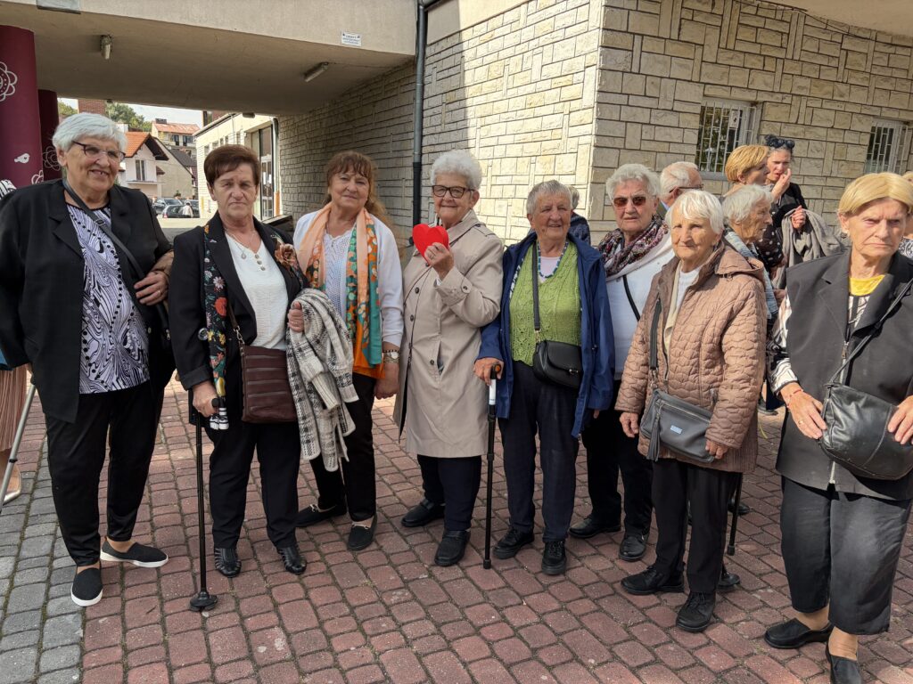 Grupka seniorek uśmiechnięta stoi w półkole, seniorka w środku trzyma drewniane czerwone serce